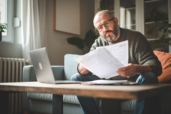 Mature man working from a couch while holding papers in his left hand with a laptop on a coffee table.