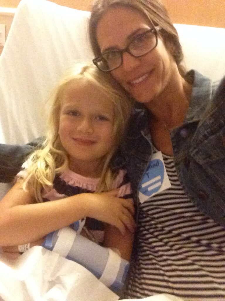 A grainy photo of a young blonde girl with her left arm protected. Her mom sits in a hospital bed with her arm around her daughter.
