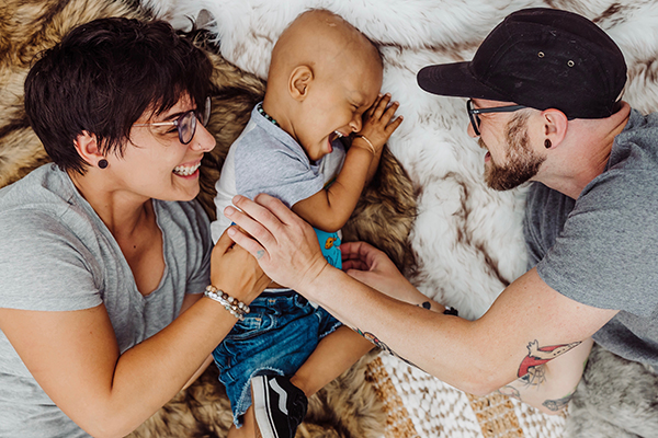 A young toddler with a bald head from chemo laughs as his mom and dad have their hands on his stomach.