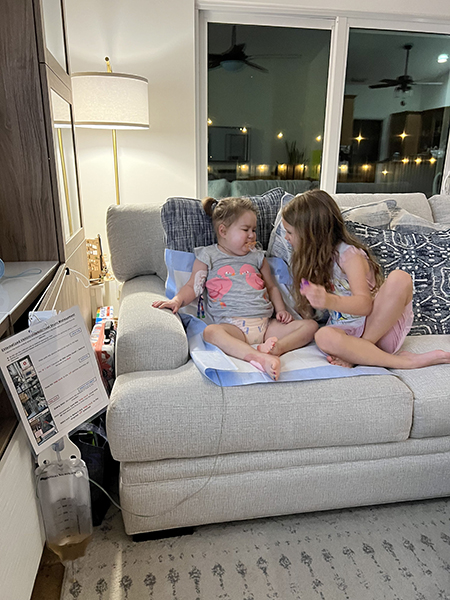 A 4-year-old girl, who has medulloblastoma, sits on a couch with her older sister. She has a pee pad under her and is hooked up to an IV.