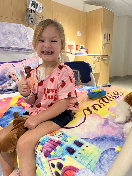A 4-year-old girl, who has medulloblastoma, has a big smile and gives a thumbs up while wearing a love bug t-shirt. She sits on top of a blanket on her hospital bed.