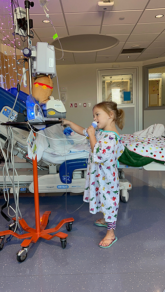 A 4-year-old girl, who has medulloblastoma, in a hospital gown, an IV, and flip flops sings with a karaoke microphone.
