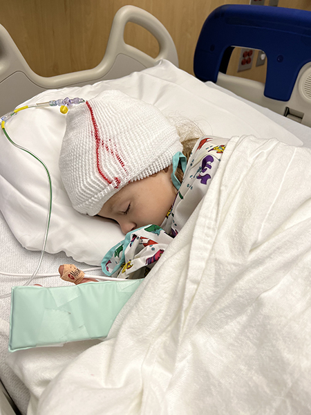 A 4-year-old girl, who has medulloblastoma, sleeps in a hospital med with white bandage and drains coming from her head and a hospital blanket over her.