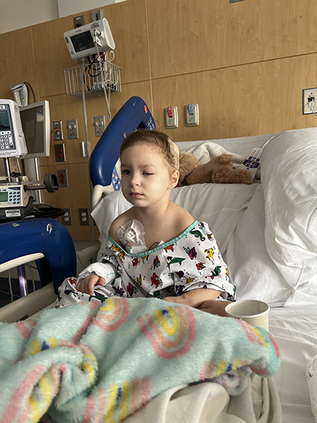 A 4-year-old girl, who has medulloblastoma and a swollen eye, sits up in a hospital bed. She has a bandage on her head, and you can see a chest port peeking out from her hospital gown.
