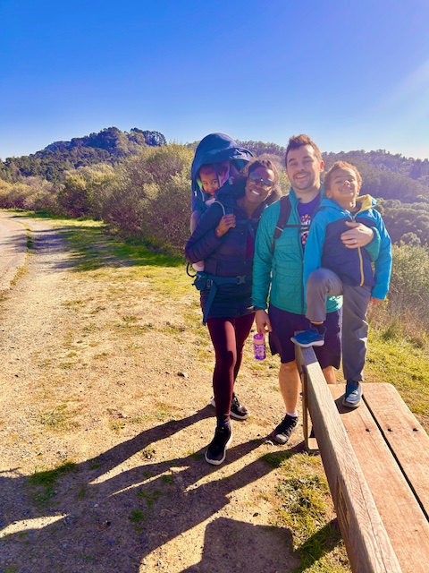 A family pauses during a hike in the California hills. A young daughter sits in a backpack carrier on her mom, while the dad has his arm wrapped around his young son standing on a bench.