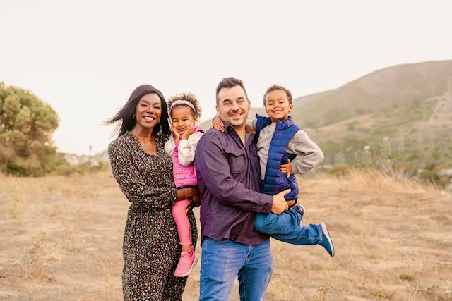 A family consisting of a mom in a black spotted dress, a young daughter in a pink jacket and pants, the dad in a purple shirt and blue jeans, and the young son in blue jeans and a blue vest jacket smile for the camera in the California hills.
