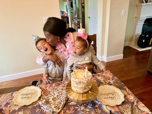 A mom kisses her young son on the cheek while holding her young daughter in her other arm. There is a birthday cake and plates in front of her.