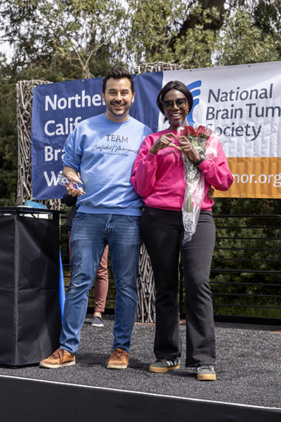A man in a blue Team Confidence Awareness sweatshirt and jeans stands on stage at the Northern California Brain Tumor Walk alongside a female survivor in a matching pink sweatshirt.