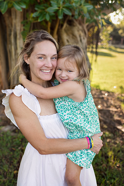 A mom in a white dress holds her 4-year-old daughter, who has medulloblastoma, wearing a green and white dress. Both are smiling at the camera.