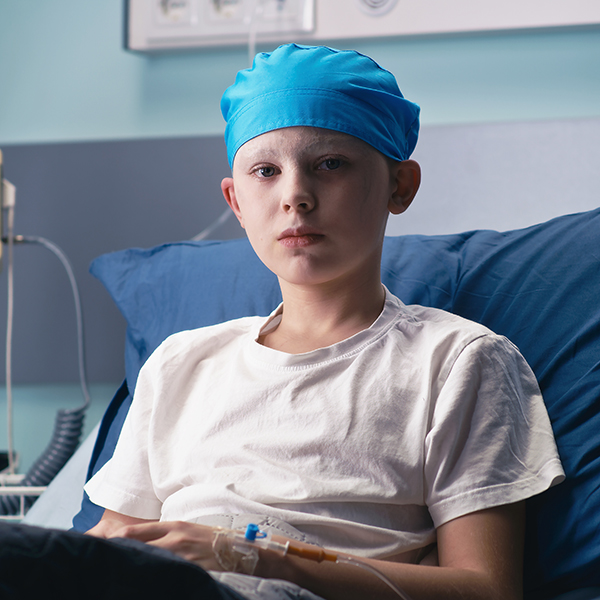 Boy in hat with medulloblastoma looking at camera while lying on bed in ward of modern cancer hospital